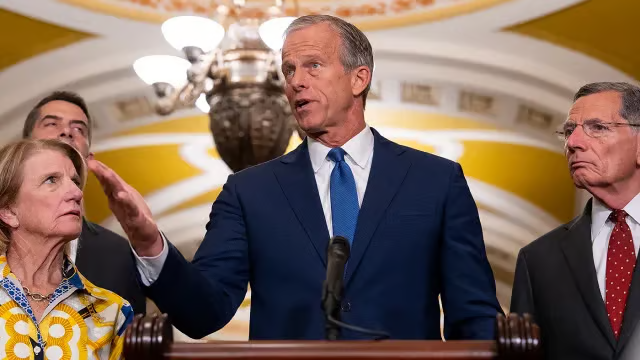 A man in a blue suit speaks at a podium with a microphone, gesturing with his hand while standing between two officials. A woman in a yellow patterned jacket is on the left and a man in a dark suit with a red tie is on the right, in an ornate room with a chandelier.